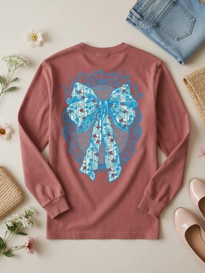 A dusty rose long-sleeve t-shirt with a large blue bow graphic on the back, styled in a flat lay with jeans, shoes, and flowers on a light beige background.